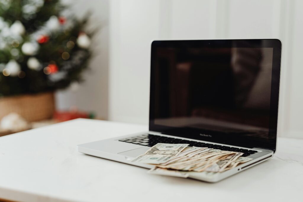 pexels photo 5942603 5942603 A laptop on a desk covered with cash, featuring a Christmas tree in the background.
