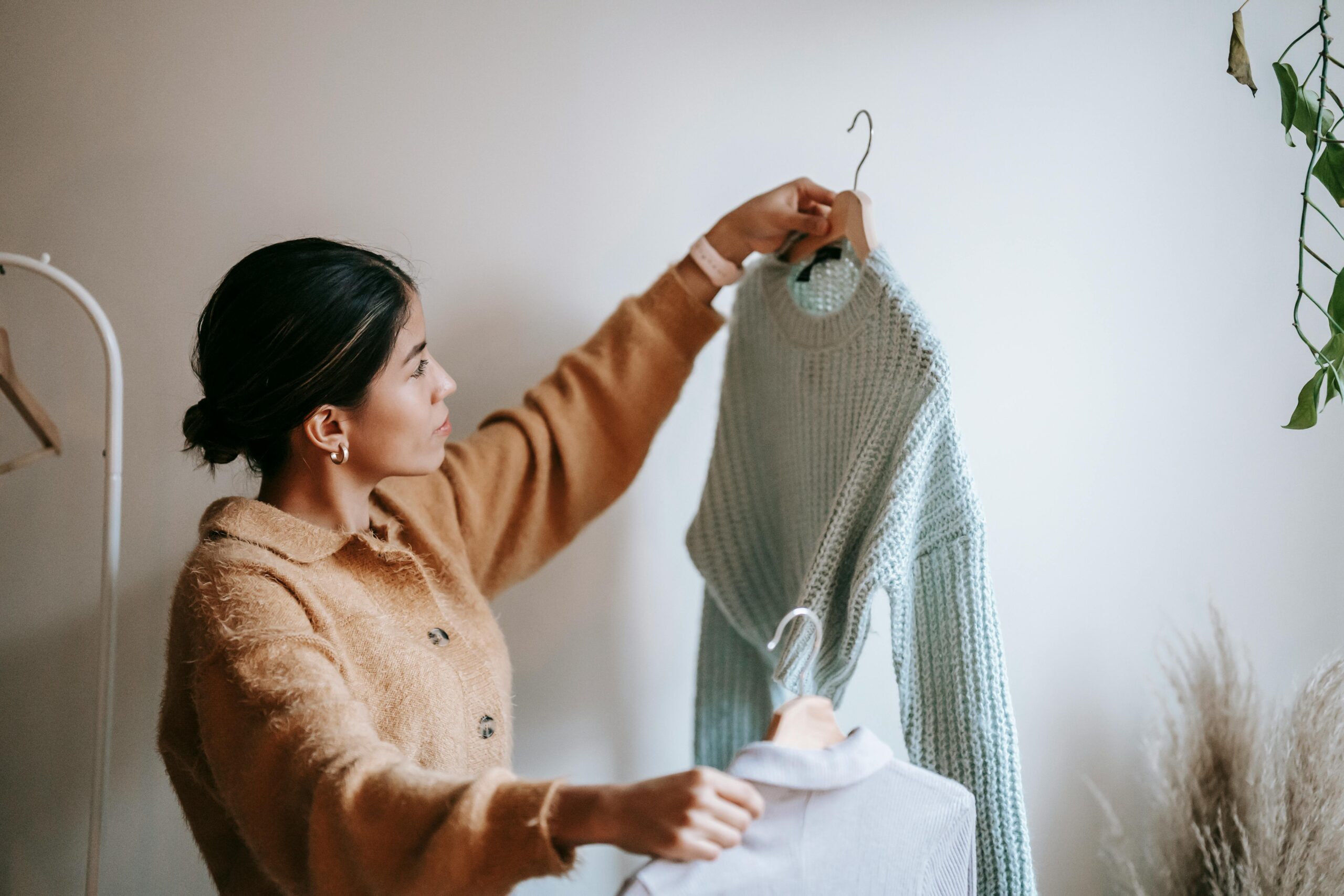 Side view of young pensive female in trendy cardigan looking at knitted sweater and shirt on hangers in cloakroom